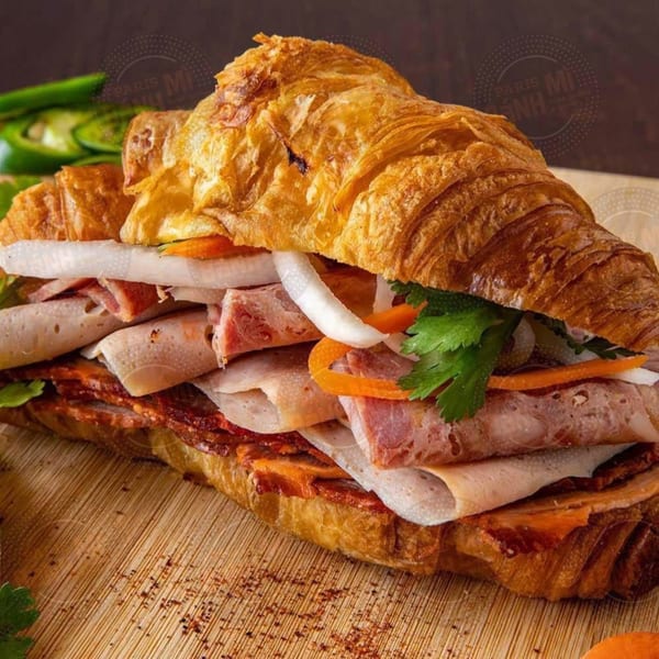 A Paris Banh Mi croissant sandwich filled with meats, pickled vegetables, and cilantro on a wooden board.