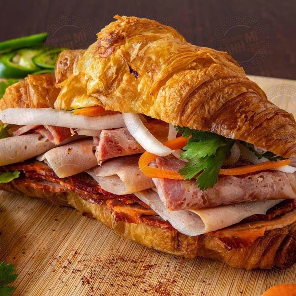 A Paris Banh Mi croissant sandwich filled with meats, pickled vegetables, and cilantro on a wooden board.