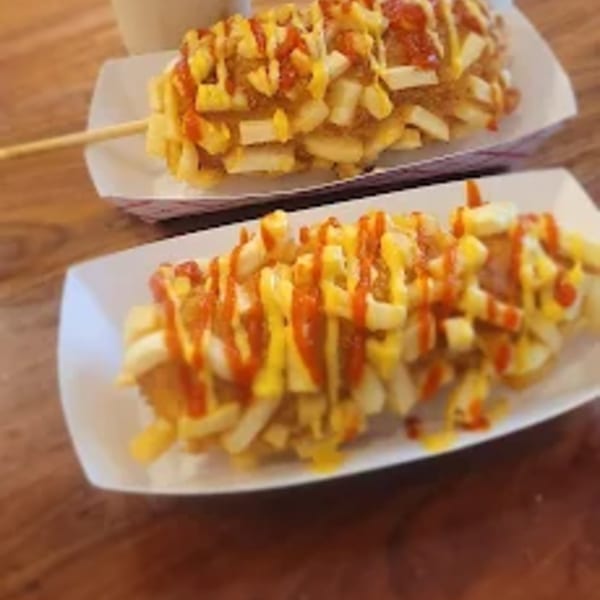 Two Korean corn dogs, covered in french fries, ketchup, and mustard, on white trays.