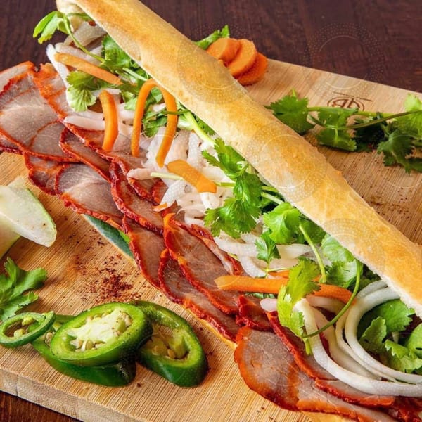 A Vietnamese banh mi sandwich with sliced pork, cilantro, carrots, and jalapenos on a wooden board.